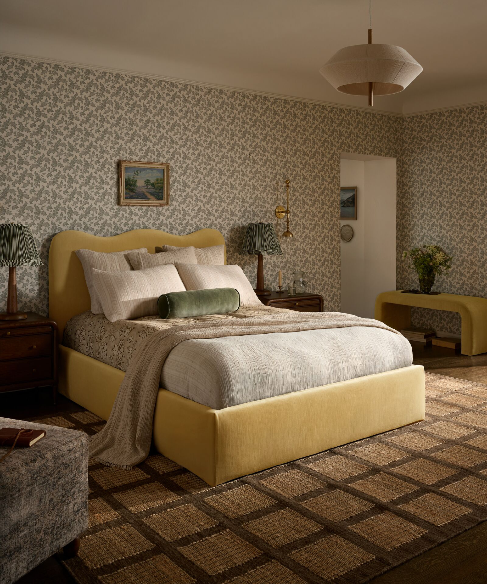 a bedroom with a yellow fabric bed, several pillows, a checkerboard-like rug, and a repetitive floral wallpaper