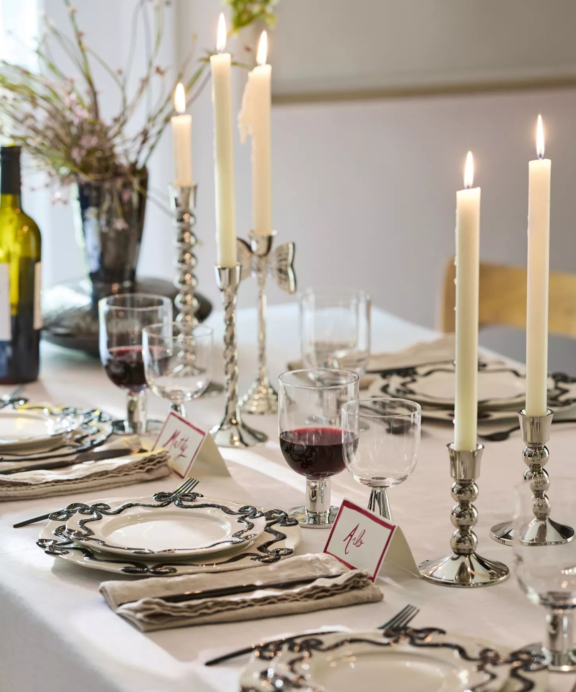 table set with Anthropologie Benedita Bow dinnerware collection, silver candlesticks, a bottle of wine, wine glasses, silver cutlery, name tags, and a vase of flowers