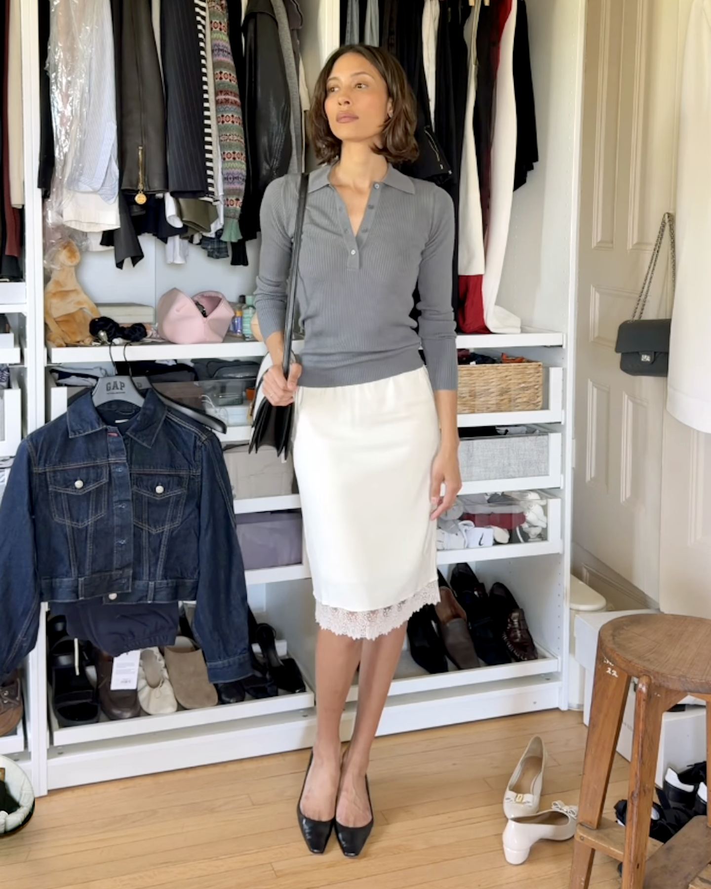 style influencer Tylynn poses in front of her closet wearing a gray long-sleeve polo shirt, a shoulder bag, a white lace-trim skirt, and black kitten-heel pumps