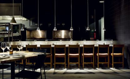 A line of wooden chairs along a counter. Four lights hang above it in an otherwise dark space