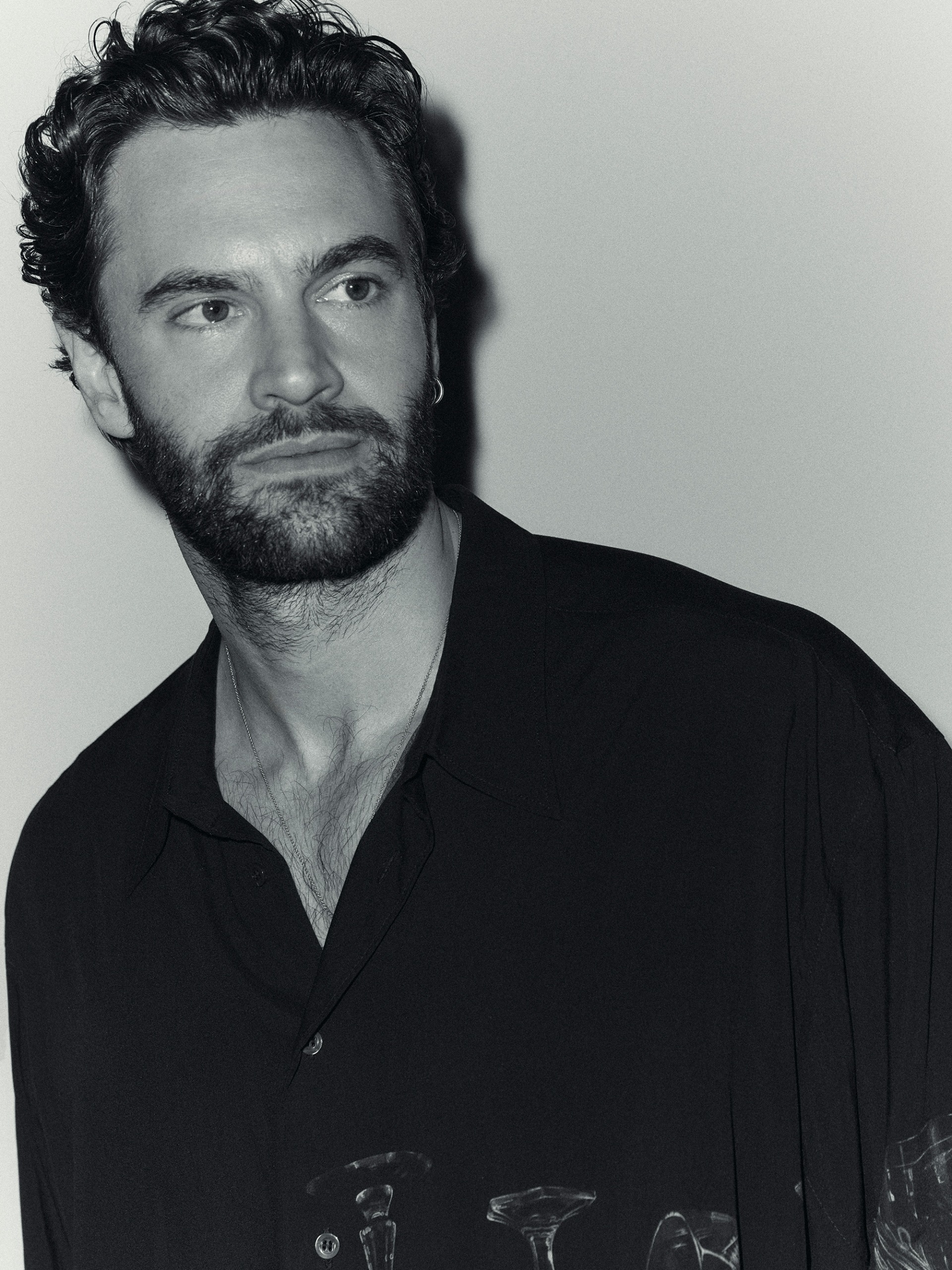 Black-and-white portrait of Tom Bateman. He is looking off-camera, wearing a button-down shirt with a print of different glassware on it.