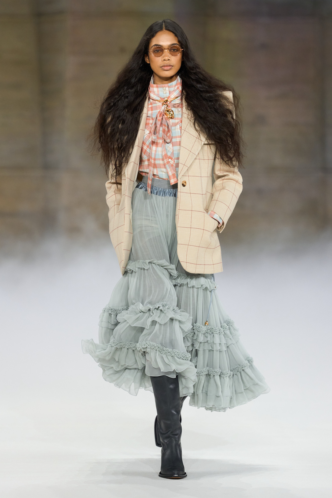 An image of a model from the autumn/winter 2026 fashion Chlo&amp;eacute; show wearing a plaid bow blouse, plaid beige blazer, dusty light blue ruffle skirt and black leather boots
