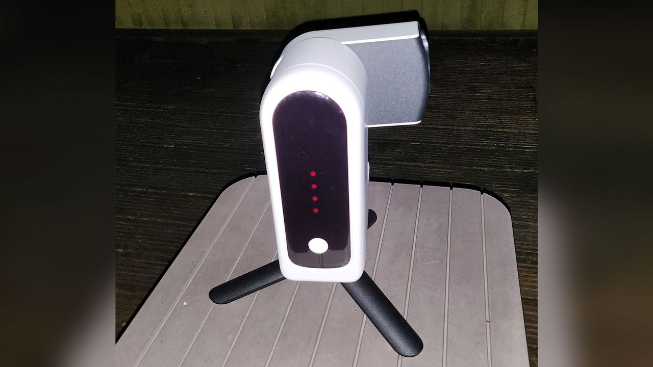 A white smart telescope with a black front is displaying red lights and a white power button is on top of three black legs on a wooden surface.