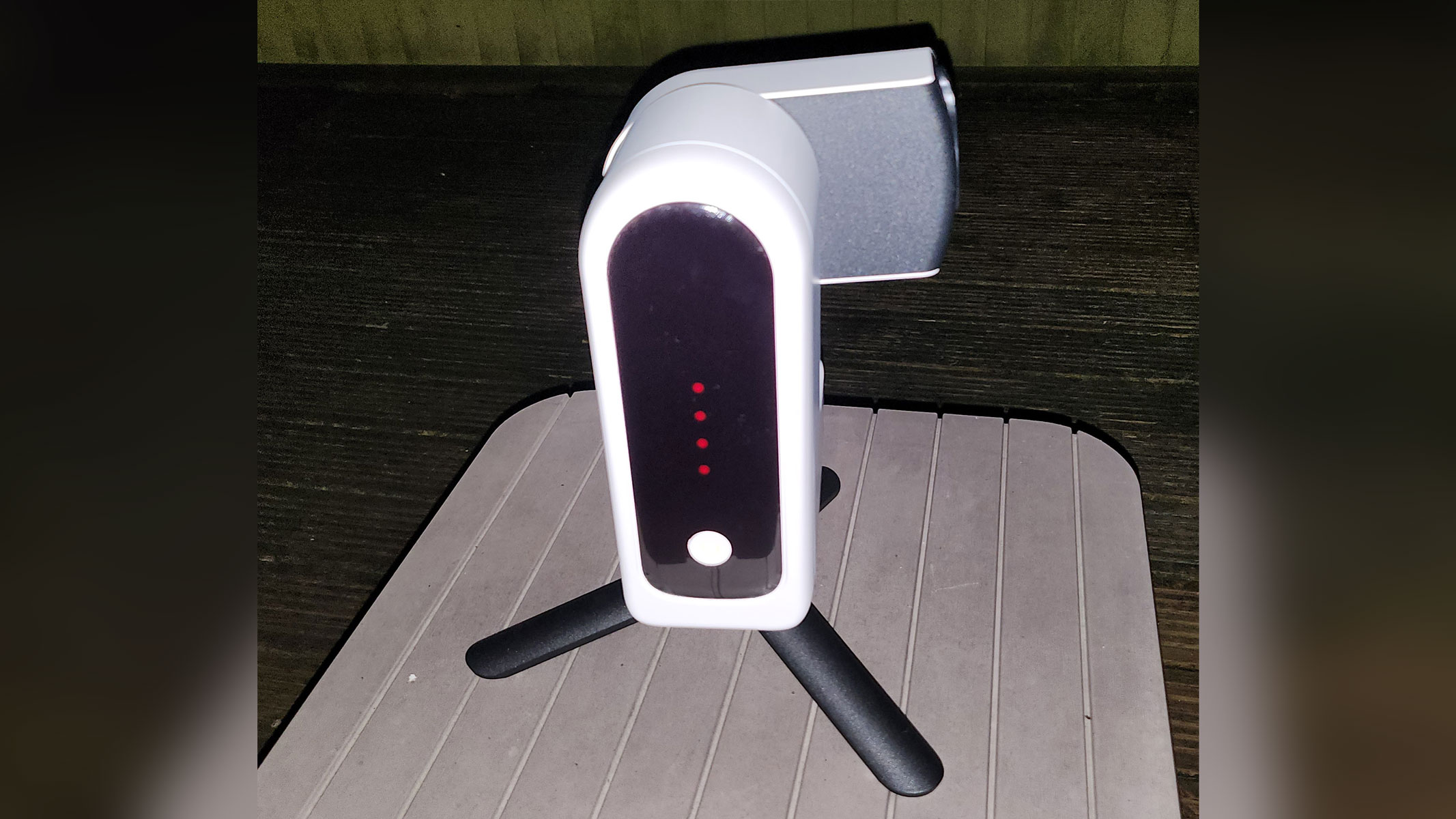 A white smart telescope with a black front is displaying red lights and a white power button is on top of three black legs on a wooden surface.