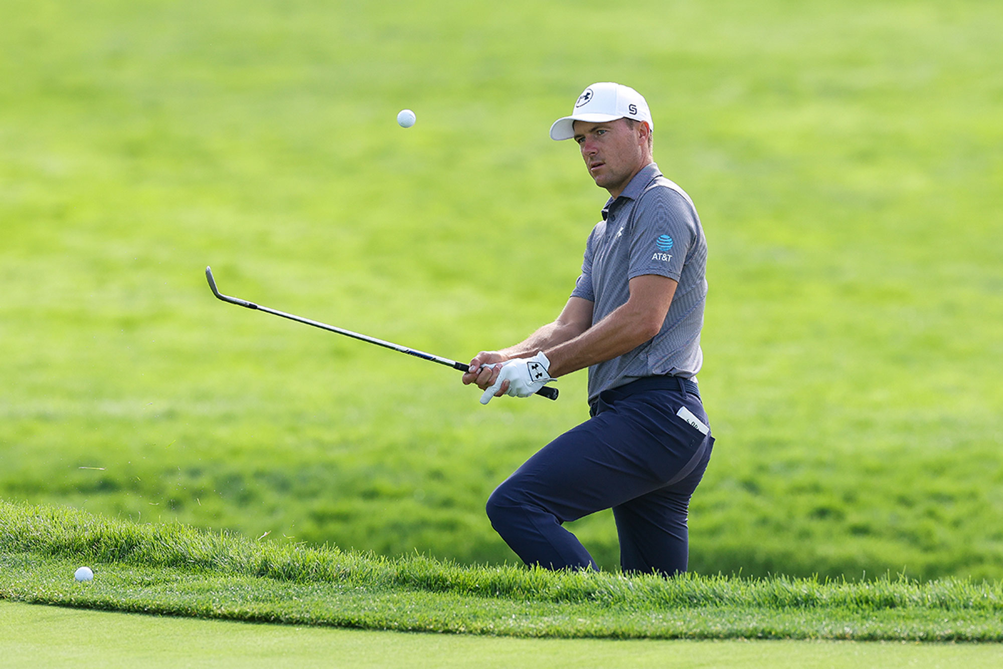 Jordan Spieth hitting a chip shot out of a bunker, with only his top half visible, and his hands down the shaft to signify a difficult lie and a tricky short game shot