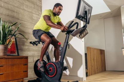 This image is of a man riding an Echelon bike in the center of the picture. The bike has a mounted screen to the right and there are weights a the back of the saddle to the left. He is riding it at home