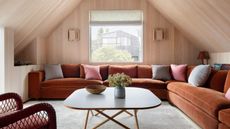conversation room in attic with timber panelled walls and ceiling, rust-colored corner sofa with cushions, square coffee table with timber legs, beige rug, and wicker armchair
