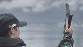 Person in baseball cap in front of lake holding up Inmarsat IsatPhone 2