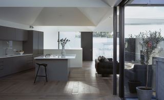 An open plan kitchen with wooden floors, gray cabinets, white ceramic top island with grey base and a structural designed white ceiling. On the right of the kitchen is a sofa