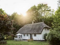 An idyllic view of the Vipp farmhouse immersed in nature with the sun setting behind the trees. The house features white plastered walls and a thatched roof, outside is a sunshade and chairs