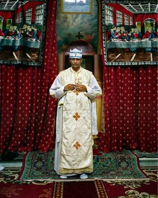 Priest in white and gold robes pictured against red and gold curtains and standing on a red Persian-like rug.