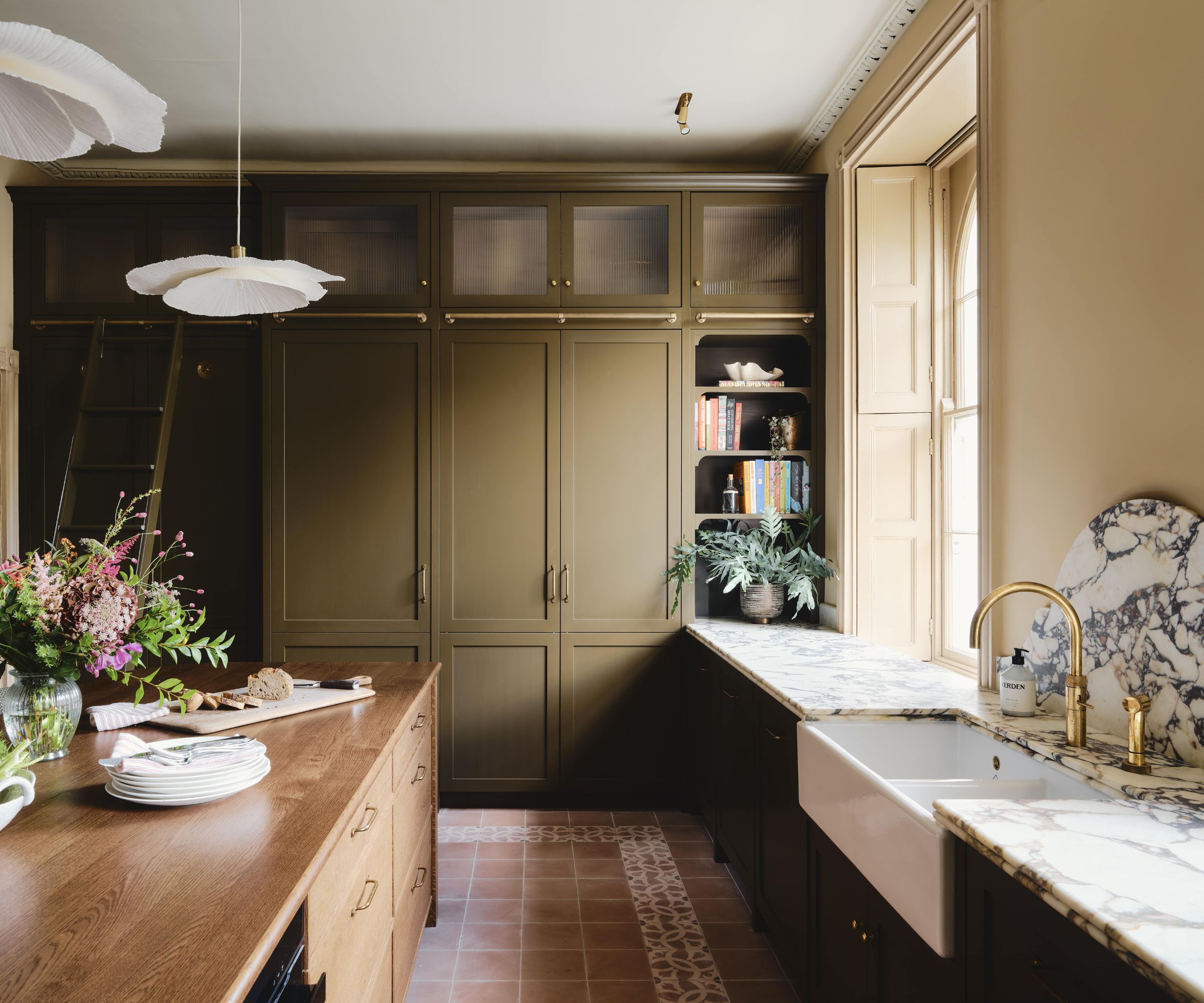A warm, country kitchen with green cabinets, a wooden island, marble countertops, and a porcelain sink