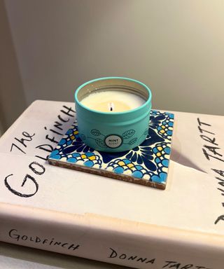 A lit candle in a green tin on a blue white and yellow patterned tile on top of a white hardback copy of The Goldfinch book against a white wall