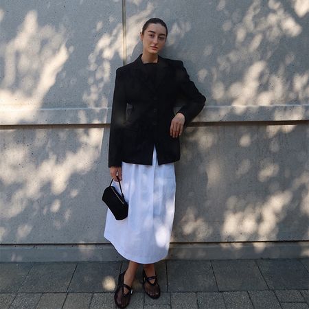 dominique wears black blazer, white skirt, black mesh flats, and black handbag while leaning against a wall