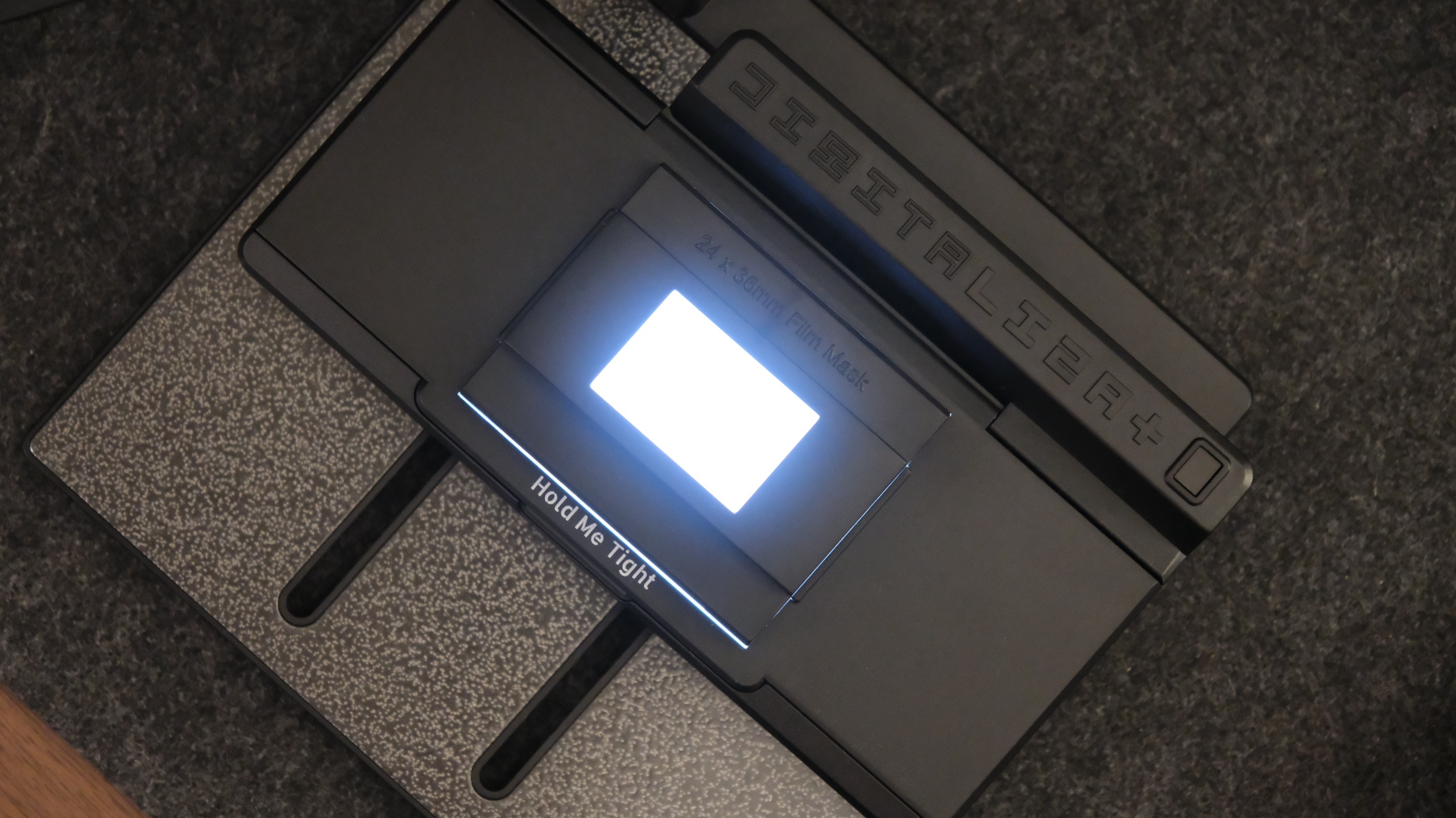 An overhead view of a film scanning base plate emitting a bright white rectangular glow, featuring an attached mask labeled "24 x 36mm Film Mask" and the text "Hold Me Tight".