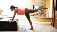 Woman exercising at home. She has both hands on a pouffe, and one leg raised behind her. She wears three-quarter length gray leggings and a pink vest top.