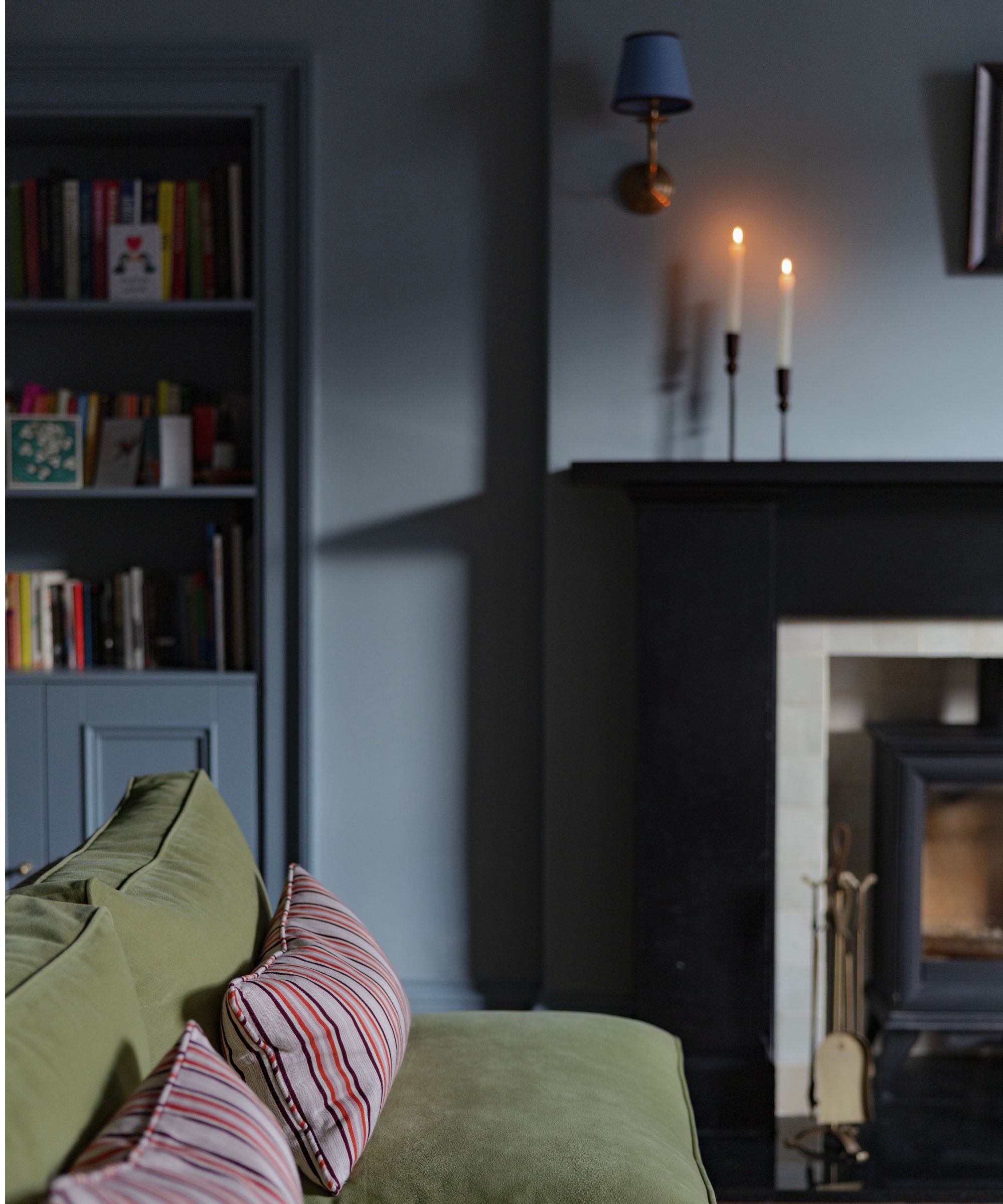 Living room with deep blue-gray walls, built-in bookshelves, a green sofa with striped cushions, and a black fireplace illuminated by candlelight.