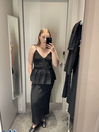 Woman in dressing room wears black top and black plisse skirt and ballet flats