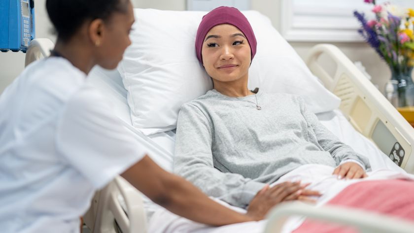 A female patient is shown sat up in a hospital bed smiling at a nurse who has their hand placed on theirs. The patient is wearing a head scarf. 