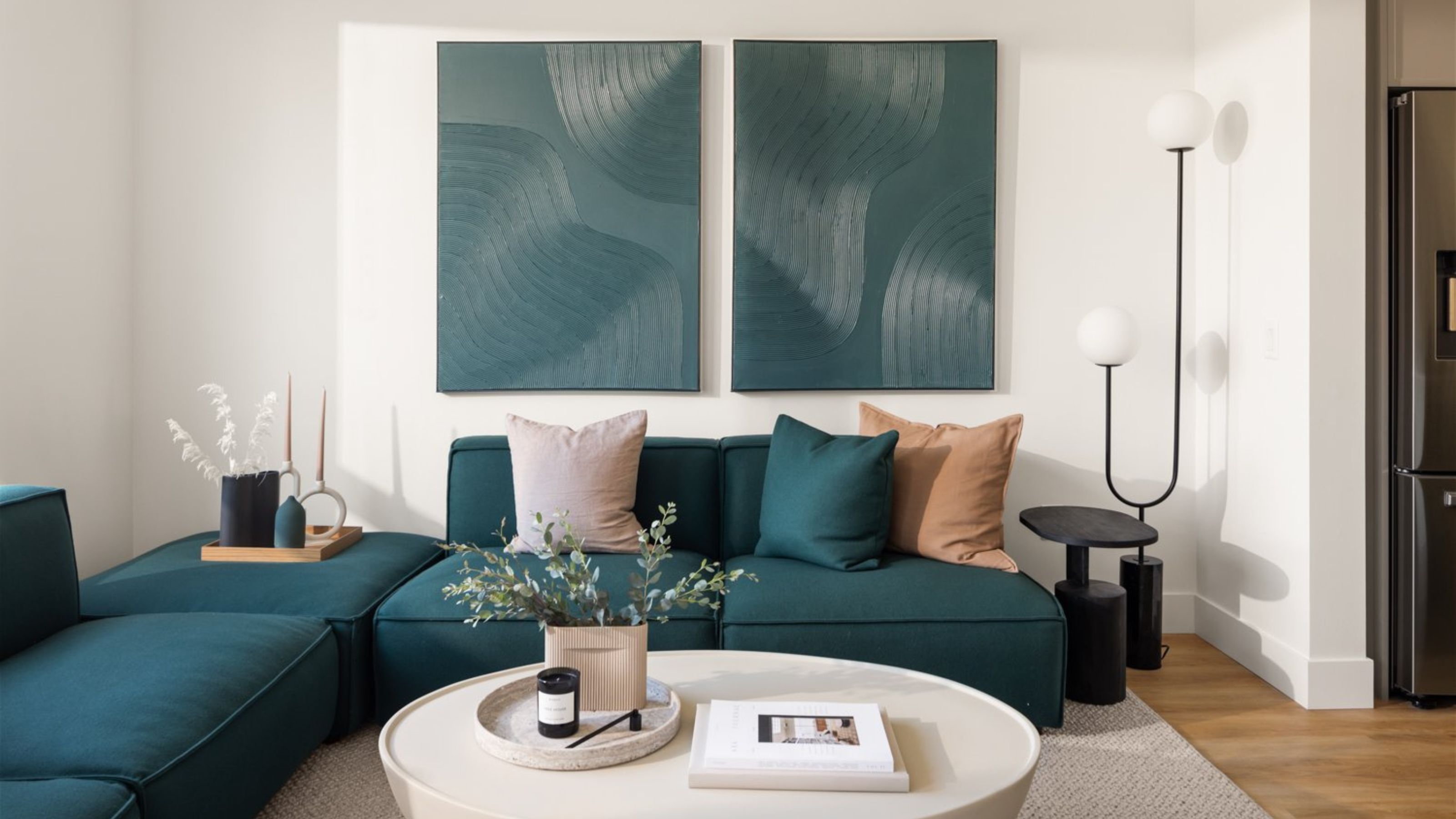 living room with white walls, a teal modular sofa with teal, cream, and tan cushions, two teal artwork on the wall above, a black side table next to a large orb-style floor lamp, a white round coffee table in front of the sofa on a white rug