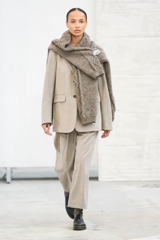 Skall Studio fall/winter 2024 model wearing a beige suit, a cable-knit sweater over her shoulders, and black combat boots.