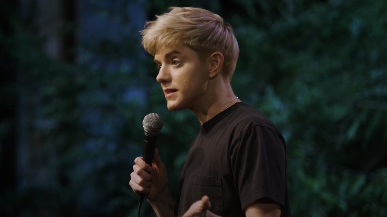 Mae Martin holding a mic onstage in their standup SAP.