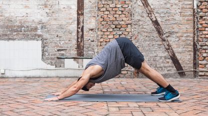 a man wearing a grey tshirt and shorts performing a downward facing dog yoga pose on a grey mat outside on brick floor and exposed brick wall behind him.