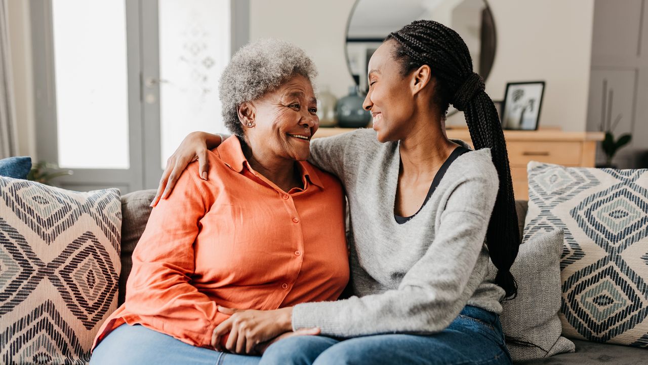 An adult daughter has her arm around her mother as they smile at each other while sitting on the sofa.