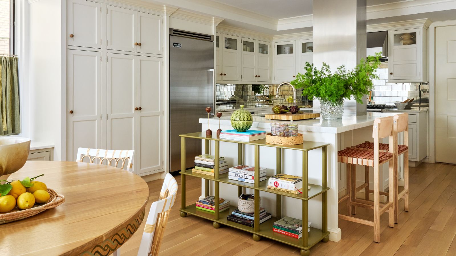 Contemporary kitchen with white cabinetry, marble-topped island and oak bar stools, softened by a freestanding Wilmington Console Table by Alfred Newall displaying books and collected objects.