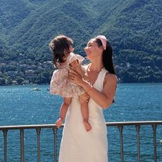 Italian woman wearing white slip dress, holding child, standing in front of Lake Como.