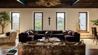Image of a large, luxurious, and cozy living room with a maximalist scheme. It features a patterned sofa, a large purple sofa, four windows across the back wall, and two potted trees.