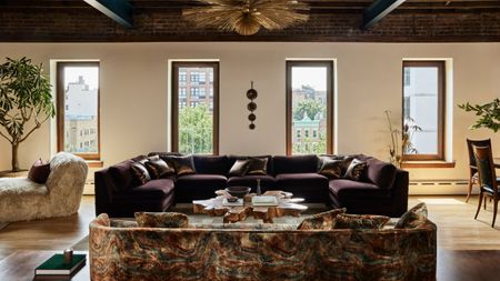 Image of a large, luxurious, and cozy living room with a maximalist scheme. It features a patterned sofa, a large purple sofa, four windows across the back wall, and two potted trees.