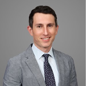 a headshot photo of Ian Williams: a man with short dark hair against a gray background in a gray suit and tie, looking at the camera
