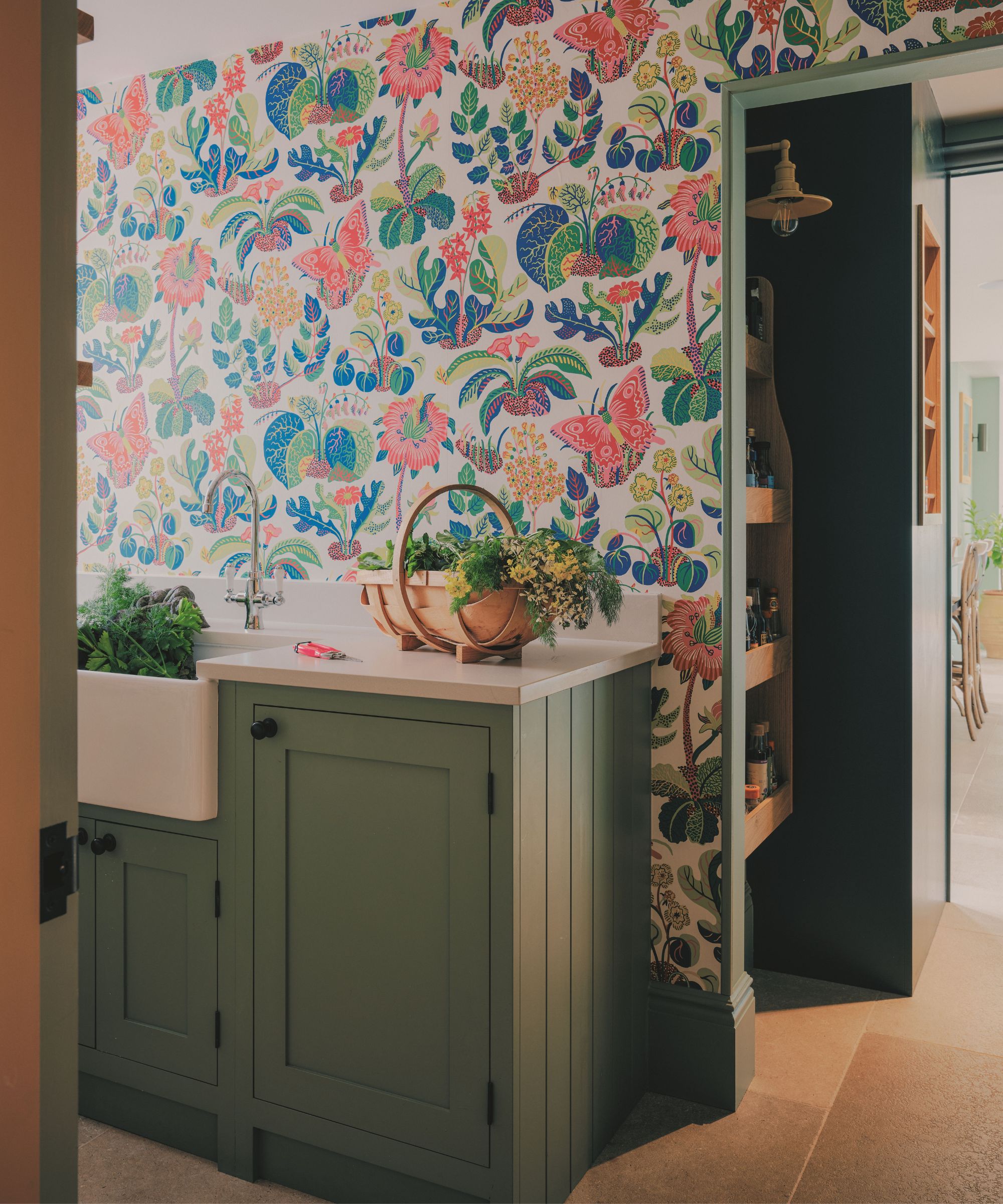 Utility area featuring colorful floral and butterfly wallpaper, sage green cabinetry, a white Belfast sink, and a wooden trug filled with fresh herbs.