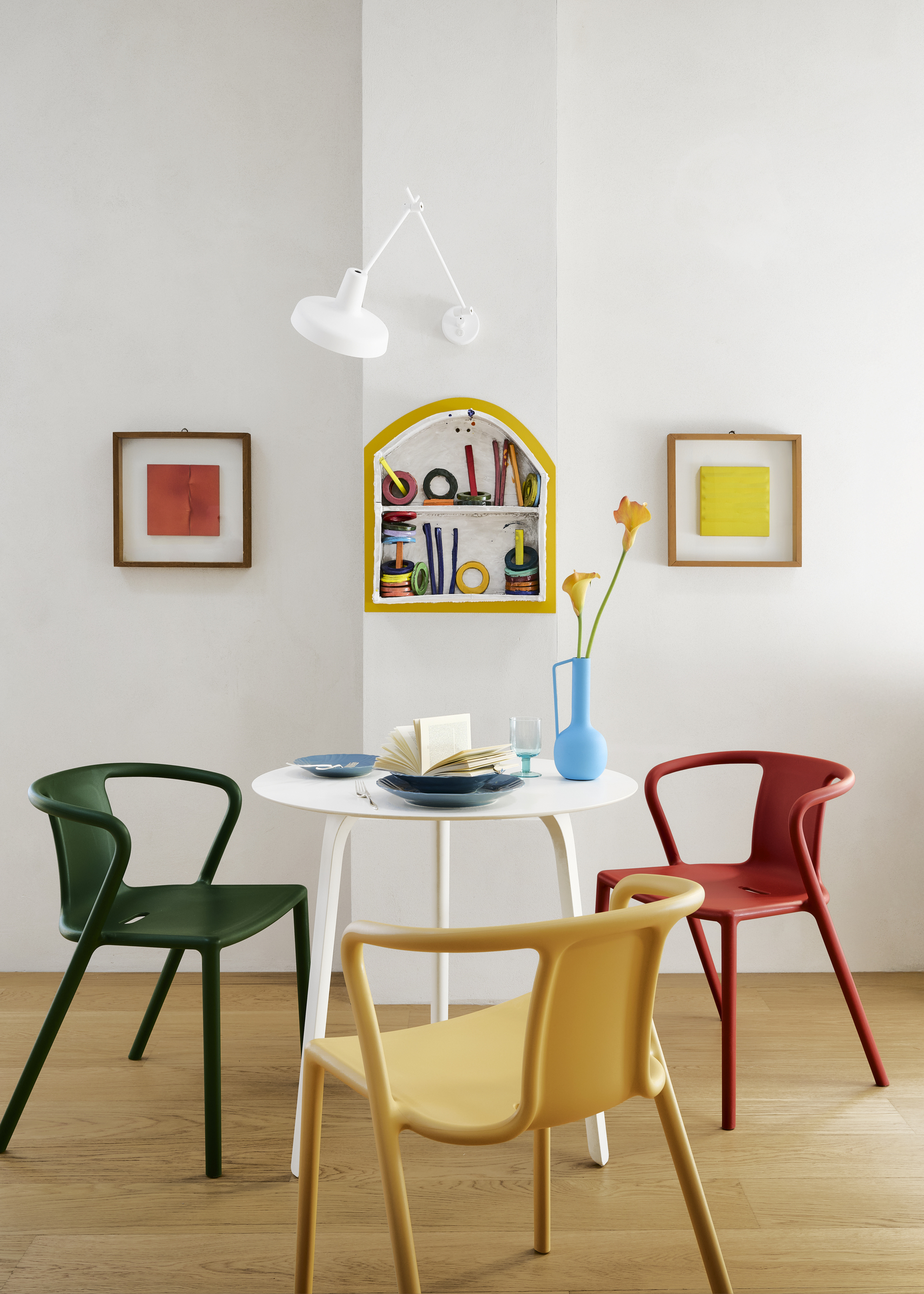 A white dining room, a wall lamp, two framed prints, a white dining table with green, red, and yellow chairs