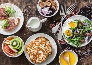 Greek yogurt with whole grain cereals and berry sauce, pancakes, arugula, cherry tomatoes, boiled eggs salad, kiwi, apples fruit, salami and cream cheese sandwiches on a wooden background, top view. Flat lay breakfast table