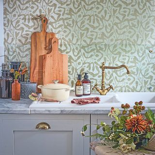 Kitchen sink area with green wallpaper behind it, brass taps, and lots of kitchen accessories