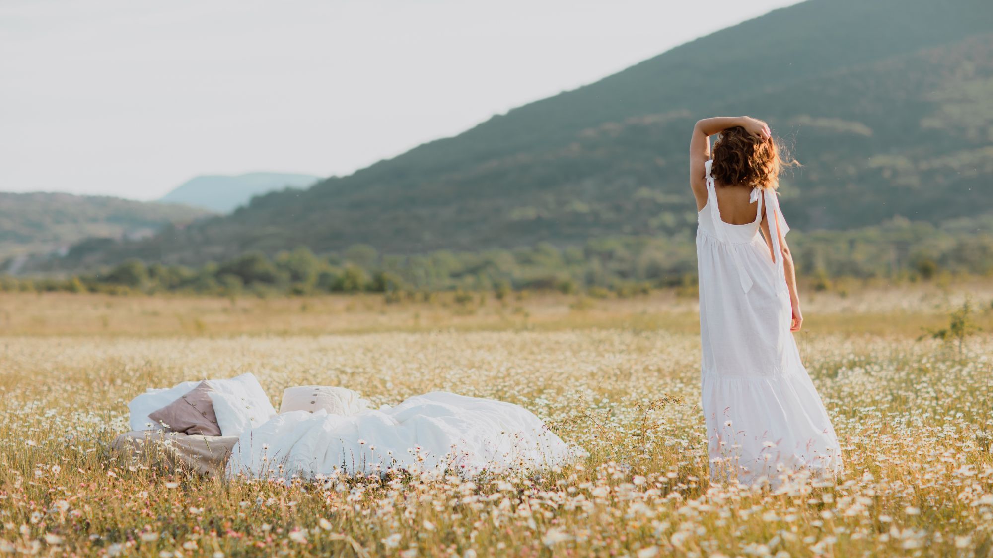 Young cute curly woman in white dress standing near soft linen bed in summer chamomile field. Summer vacations inspiration