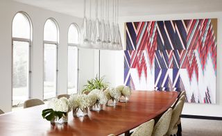Dining room with large wooden table, vases of white flowers, white and glass lighting feature and patterned chairs.