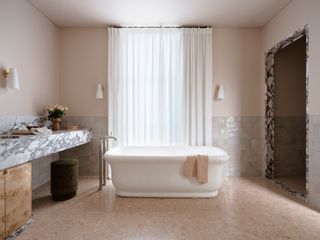 Neutral bathroom with pale pink walls and off-white zellige tiles, white freestanding bath, marble countertop and marble trim around base of vanity unit and entrance to walk-in shower