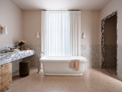 Neutral bathroom with pale pink walls and off-white zellige tiles, white freestanding bath, marble countertop and marble trim around base of vanity unit and entrance to walk-in shower