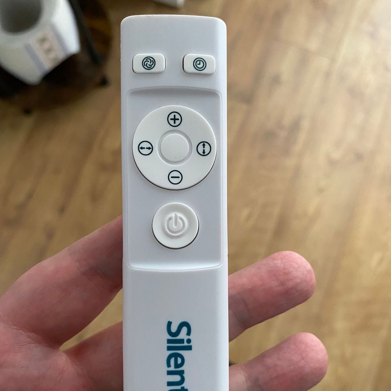 A hand holding the white remote control of the Silentnight Airmax Pro360 Pedestal Fan, with wood-look floor in the background