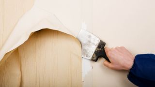 Wallpaper being removed from a wall with a metal hand tool, representing an article about how to remove wallpaper