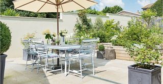 summer garden with a bright tiled patio with a wining table with a parasol surrounded by potted plants partially shaded to avoid sunburnt plants