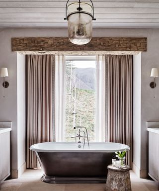 Bathroom with paneled wooden ceiling, looking onto metal bathtub beside window, metal side table, stone flooring, glass and metal lantern style pendant light,