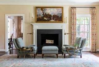fireplace with antique landscape painting above and two blue armchairs and ottoman on patterned rug with pale walls and patterned curtains