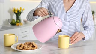 Pink kettle being poured into a yellow mug next to biscuits