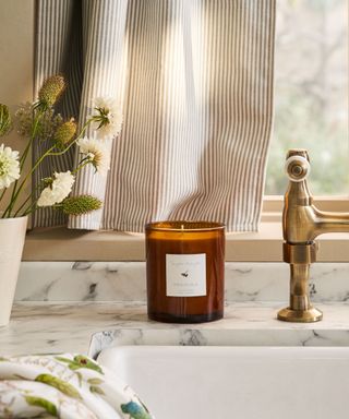 A brown glass jar candle on a marble kitchen counter beside an inset sink with gold taps. The edge of a white vase with blooming white flowers on the left side.