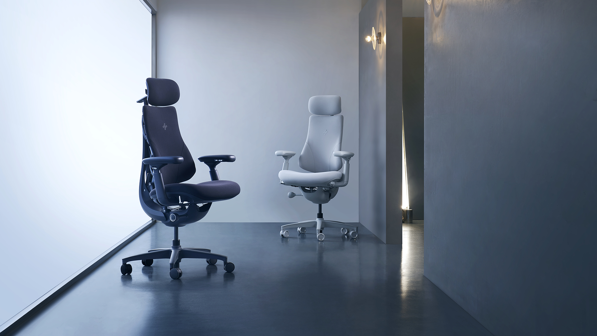 Two ergonomic office chairs, one dark and one light, displayed in a minimalist showroom with cool lighting and polished floors.