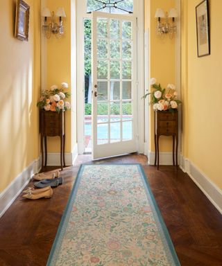 An entryway with yellow walls, a white door, two dark wooden tables with flowers on top, and a dark wooden floor with a long blue floral runner rug on it